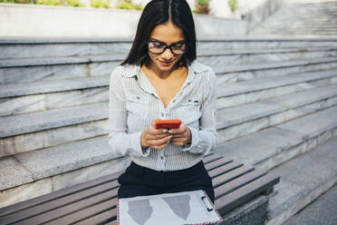 Junge Geschäftsfrau, die auf einer Bank sitzt und ihr Mobiltelefon überprüft - OYF00036