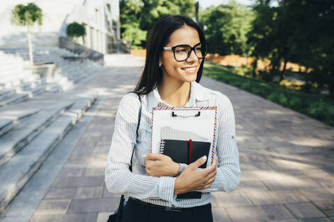Lächelnde junge Frau beim Spaziergang mit Papieren im Freien, lizenzfreies Stockfoto