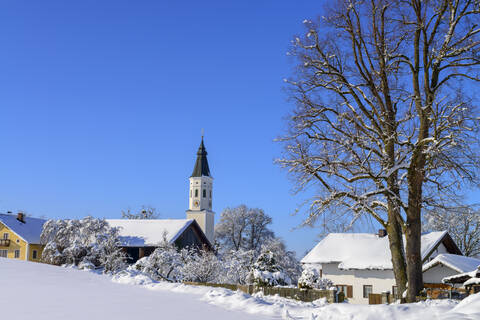Kirche in schneebedeckter Landschaft und kahler Baum vor blauem Himmel, Bayern, Deutschland, lizenzfreies Stockfoto