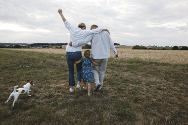 Rückansicht einer Familie mit Hund beim Spaziergang auf einer Wiese - KMKF01052