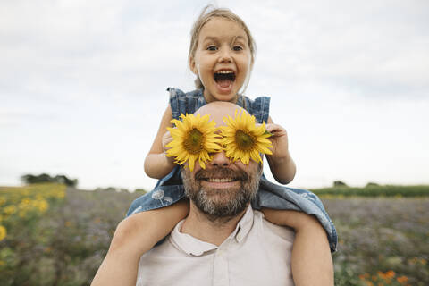 Sonnenblumen bedecken die Augen eines verspielten Mannes mit Tochter auf einem Feld, lizenzfreies Stockfoto