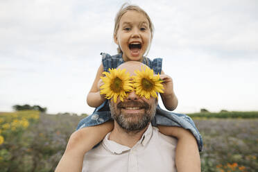 Sonnenblumen bedecken die Augen eines verspielten Mannes mit Tochter auf einem Feld - KMKF01063