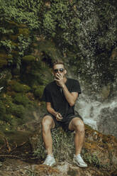 Porträt eines coolen jungen Mannes an einem Wasserfall, der einen Joint Marihuana raucht - ACPF00629