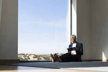Älterer Geschäftsmann sitzt am Panoramafenster und benutzt ein Tablet - SBOF02003