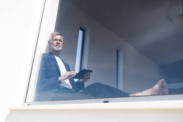 Senior businessman sitting at panorama window using tablet - SBOF02004