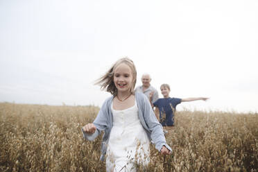 Porträt eines glücklichen Mädchens, das in einem Haferfeld läuft, während Bruder und Großvater ihr folgen - EYAF00414