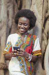 Lächelnde junge Frau lehnt an einem Baumstamm und benutzt ihr Smartphone - DLTSF00019