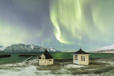 Das Nordlicht (Aurora Borealis) und die Sterne beleuchten die Holzhütten am eisigen Meer, Djupvik, Lyngen Alps, Troms, Norwegen, Skandinavien, Europa - RHPLF07946