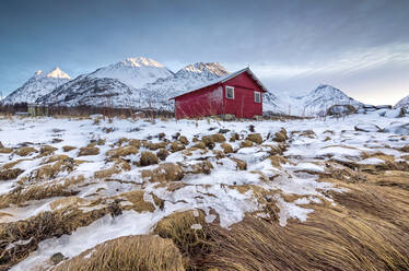 Holzhütte umrahmt von mit Gras und Eis bedeckten Felsen mit schneebedeckten Gipfeln im Hintergrund, Svensby, Lyngen Alps, Troms, Norwegen, Skandinavien, Europa - RHPLF07953