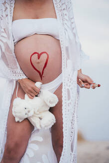 Schwangere Frau mit einem gezeichneten Herz auf dem Bauch, die einen Teddybär hält - JCMF00140