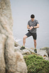 Trail runner standing in coastal landscape looking at his watch, Ferrol, Spain - RAEF02284