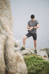 Trailrunner steht in einer Küstenlandschaft und schaut auf seine Uhr, Ferrol, Spanien - RAEF02284