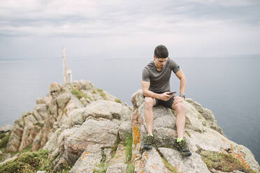 Trailrunner sitzt auf einem Felsen in der Küstenlandschaft und schaut auf sein Handy, Ferrol, Spanien - RAEF02289