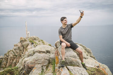 Trail runner sitting on a rock in coastal landscape taking a selfie, Ferrol, Spain - RAEF02290