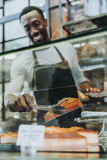 Lächelnder Mann, der in einer Bäckerei arbeitet und ein Stück Kuchen auf einen Teller legt - JCMF00152