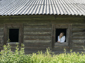 Frau schaut aus dem Fenster eines alten Holzhauses im Dorf - AHSF00779