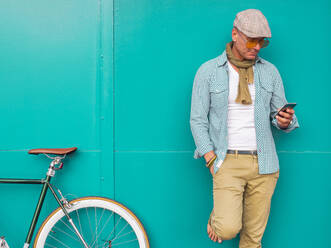 Mann mit Fixie-Fahrrad steht vor grüner Wand und schaut auf sein Handy - DLTSF00051