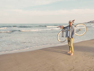 Barfuss stehender Mann mit Fixie-Fahrrad auf der Schulter am Strand - DLTS00054