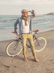 Barfußmann mit Fixie-Fahrrad am Strand stehend und in die Ferne blickend - DLTSF00055