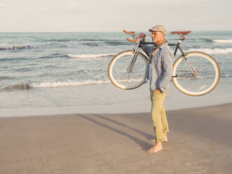 Barefoot man standing with Fixie bike on his shoulder on the beach - DLTSF00056