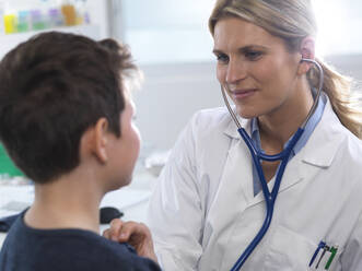 Female doctor listening to a boys heart beat using a stethoscope during a health check in a clinic - ABRF00623