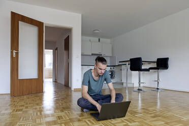 Junger Mann sitzt in einer leeren Wohnung und benutzt seinen Laptop - FLLF00282