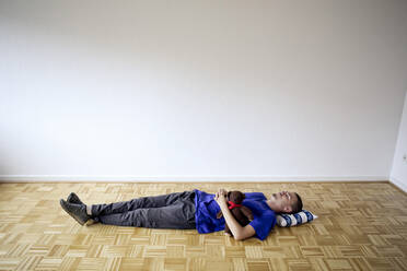 Junger Mann liegt in einer leeren Wohnung auf dem Boden und umarmt einen Teddybär - FLLF00287