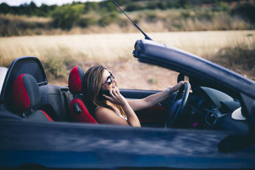 Junge Frau mit Sonnenbrille, die in ihrem Cabrio ihr Smartphone benutzt - OCMF00605