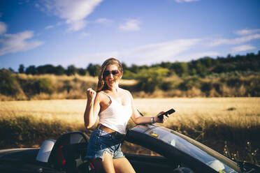 Frau steht in ihrem Cabrio, hält Smartphone, freut sich über gute Nachrichten - OCMF00610