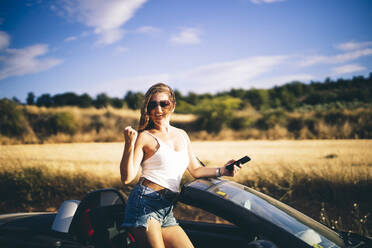 Woman standing in her convertible, holding smartphone, happy about good news - OCMF00610