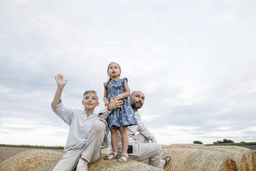 Father and his children sitting on hay bales, watching sky - KMKF01080