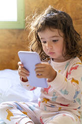 Niedliches kleines Mädchen im Schlafanzug im Bett, das ein Mobiltelefon benutzt - GEMF03124