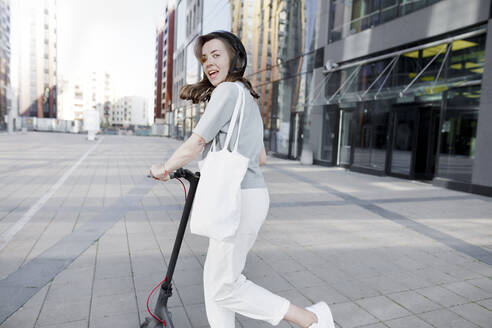 Frau mit E-Roller und Helm, moderne Bürogebäude im Hintergrund - KMKF01090