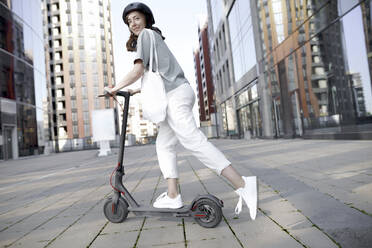 Frau mit E-Roller und Helm, moderne Bürogebäude im Hintergrund - KMKF01092