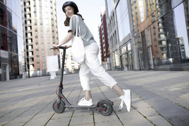 Woman with e-scooter and helmet, modern office buildings in the background - KMKF01092