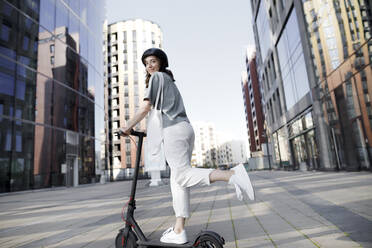 Frau mit E-Roller und Helm, moderne Bürogebäude im Hintergrund - KMKF01093