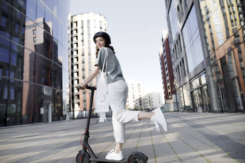 Woman with e-scooter and helmet, modern office buildings in the background - KMKF01093