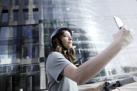 Frau mit E-Roller und Helm, die ein Smartphone benutzt, moderne Bürogebäude im Hintergrund, lizenzfreies Stockfoto