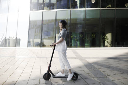 Frau mit E-Roller und Helm, moderne Bürogebäude im Hintergrund - KMKF01100