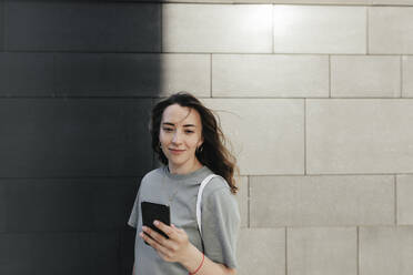 Smiling woman using smartphone, grey wall in the background - KMKF01118