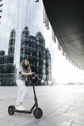 Frau mit E-Roller und Helm, moderne Bürogebäude im Hintergrund - KMKF01126