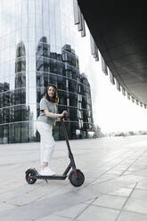 Frau mit E-Roller und Helm, moderne Bürogebäude im Hintergrund - KMKF01127