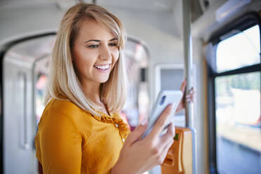 Lächelnde junge Frau, die in einer Straßenbahn ihr Smartphone benutzt - BSZF01332