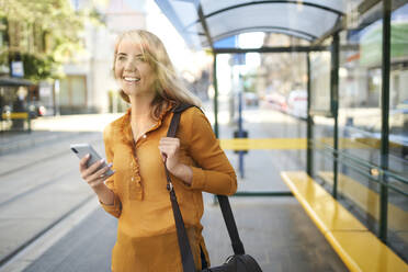 Smiling young woman with smartphone waiting for the tram at tram stop - BSZF01333