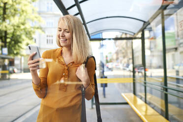 Lächelnde junge Frau mit Smartphone an der Straßenbahnhaltestelle - BSZF01335