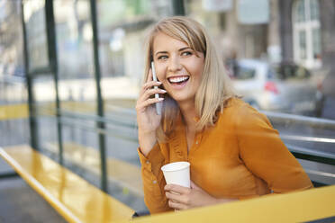 Happy young woman with smartphone and takeaway coffee at bus stop - BSZF01340