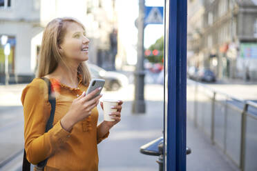 Smiling young woman with smartphone and takeaway coffee checking the schedule at bus stop - BSZF01341