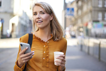 Smiling young woman with smartphone and takeaway coffee in the city - BSZF01342