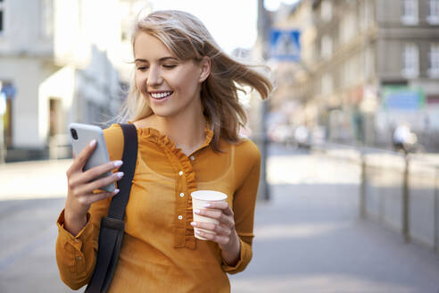 Lächelnde junge Frau mit Smartphone und Kaffee zum Mitnehmen in der Stadt - BSZF01343