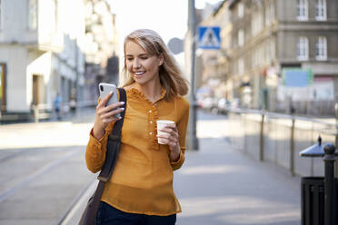 Smiling young woman with smartphone and takeaway coffee in the city - BSZF01344